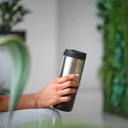 A drink bottle with a stainless steel body and a black lid, held by a hand against a green backdrop.