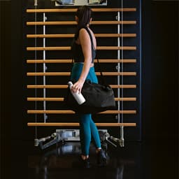 A woman in athletic wear carries a black gym bag and holds a white drink bottle, standing before a wooden gym structure.