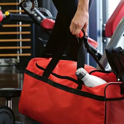 A large red gym bag with black straps, featuring a reflective strip, sits beside gym equipment. A drink bottle is visible inside.