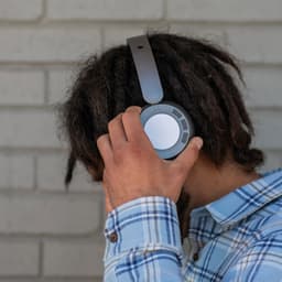 A person with dreadlocks wears grey headphones with a round earcup, against a light brick background.