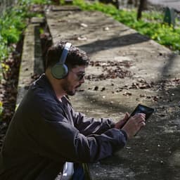 A man wearing grey headphones interacts with a tablet while sitting outdoors on a stone bench surrounded by greenery.