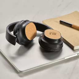 Black headphones with cork detailing, resting on a laptop next to a notebook and pen. The headphones have a sleek design.