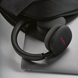 Black headphones with grey cushioning resting on a laptop, partially visible in a bag. Features a logo on the ear cup.