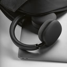 A pair of black headphones resting on a silver laptop, partially covered by a dark bag. The headphones have a sleek design.