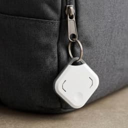 A grey fabric bag featuring a white VibeTag tracker attached to a zipper with a keyring.