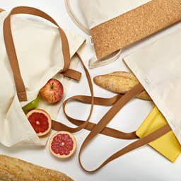 A cotton shopping bag with a cork panel, featuring brown straps and filled with fruits and baguettes.