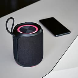 A black portable speaker with pink accents sits next to a smartphone on a white surface.
