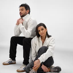 A white 1/4 zip sweatshirt is displayed on a seated man and woman, both wearing casual outfits. The sweatshirt features a logo.