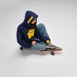 A navy oversized hooded sweatshirt is worn by a person sitting on a skateboard, paired with baggy jeans and sneakers.