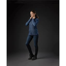 A woman stands wearing a navy thermal shell jacket with a quilted design, paired with dark jeans and boots.