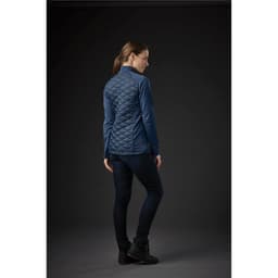 A woman wearing a blue quilted thermal shell jacket, viewed from the back, with dark jeans and footwear.