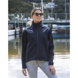 A woman wearing a navy softshell jacket, standing by the water with boats in the background. The jacket has a logo.
