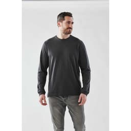 A man wearing a long sleeve tee in dark grey, paired with light grey jeans, against a plain background.