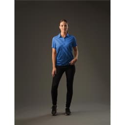 A woman stands wearing a blue Women's Reflex Polo shirt paired with black pants in a studio setting.