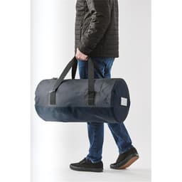 A person carrying a navy waterproof duffel bag with black straps and a branding logo.