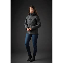 A woman stands in a black Women's Epsilon System Jacket, paired with blue jeans, against a dark background. The jacket features a logo.