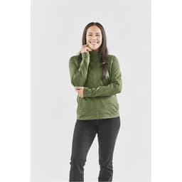 A woman stands smiling in a green performance jacket, paired with black pants. The jacket features a high collar and a logo.
