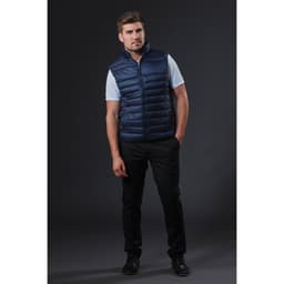A man wearing a navy puffer vest over a white shirt, paired with black trousers and shoes, standing against a dark background.