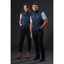 Two models wearing navy blue puffer vests, standing side by side against a dark background. The vests have a zip closure and a sleek design.