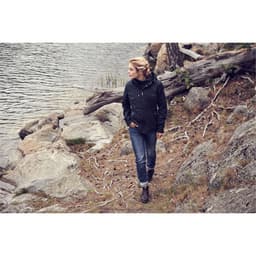 A woman walks along a rocky path by the water, wearing a black rain jacket and casual jeans.