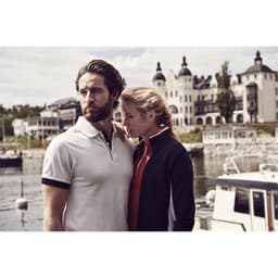 A man in a white polo shirt with black accents stands next to a woman in a dark jacket, near a waterfront.