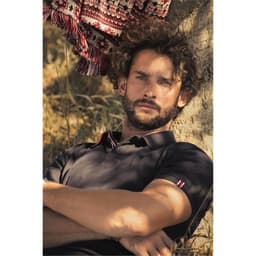 A black polo shirt worn by a man with curly hair, leaning against a tree, with a patterned fabric hanging nearby.