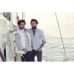 Two men posing on a sailboat, one wearing a light blue button-up shirt and the other in a white vest over a checked shirt.