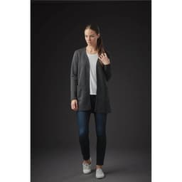 A woman wearing a grey quilted cardigan over a white top and jeans stands against a dark background.