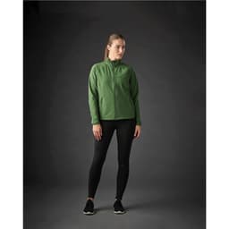 A woman stands wearing a green Women’s Kyoto Jacket paired with black leggings and shoes, with a dark background.
