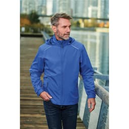 A man wearing a blue rain jacket with a hood, standing on a wooden boardwalk near water. The jacket features a logo.