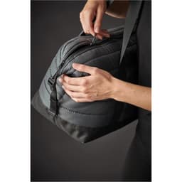 A quilted duffle bag in grey with a zippered opening, being held by hands. Features a logo on the side.