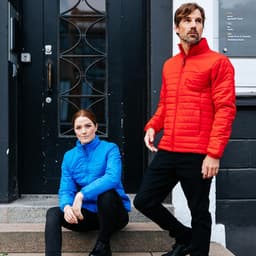 A woman in a blue quilted jacket and a man in a red quilted jacket stand on steps outside a building. Both jackets have branding.