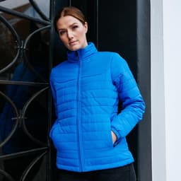 A blue women's quilted jacket with a high collar and two side pockets, modelled against a decorative door.