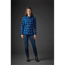 A woman stands wearing a blue and black checked button-up shirt and jeans, with a plain dark background.