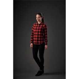 A woman stands in a black and red checked button-up shirt paired with black pants and shoes. The shirt features a logo.