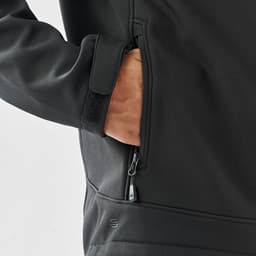 A black softshell jacket with a zippered pocket, featuring a hand reaching inside the pocket.