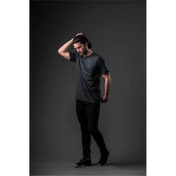 A model wearing a short-sleeve, dark grey T-shirt paired with black pants against a dark background. The shirt features a logo.