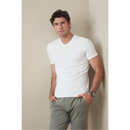 A man in a white V-neck t-shirt stands against a neutral background, hands in pockets, wearing casual pants.