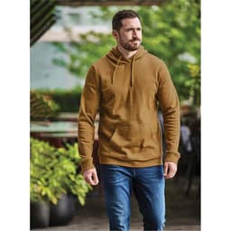 A man wearing a brown fleece pullover hoody, paired with blue jeans, walks outdoors in a leafy setting.