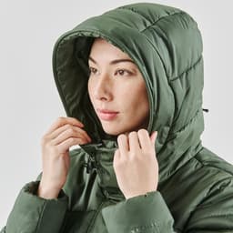 A woman wearing a green puffer jacket with a hood, adjusting it around her face. The jacket has a subtle logo.