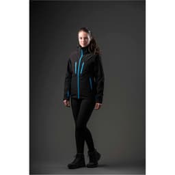 A woman wearing a black jacket with blue accents, paired with leggings and boots, standing against a dark background.