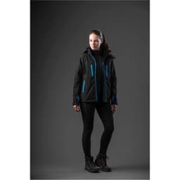 A woman in a black jacket with blue accents stands against a dark background, wearing black leggings and boots.