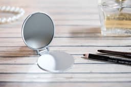 A compact silver pocket mirror sits open on a wooden surface, accompanied by two makeup pencils and a perfume bottle.