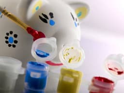 A white plaster piggy bank with painted features, surrounded by small pots of blue, yellow, red, and green paint.