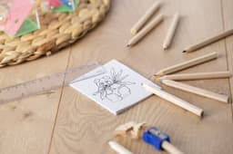 A set of coloured pencils in natural wood tones surrounds a drawing pad featuring an outline illustration.