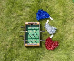 A MDF football table game in brown, featuring green pitch, with red and yellow players, and blue and silver pom-poms.
