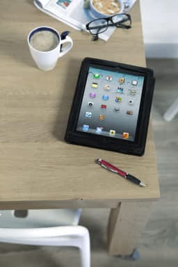 A black tablet sits on a wooden table beside a coffee cup and a red ABS ballpen, with glasses and a newspaper in the background.
