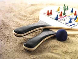 A beach ball game set featuring two paddles with colourful pegs and a blue ball, resting on sandy ground.