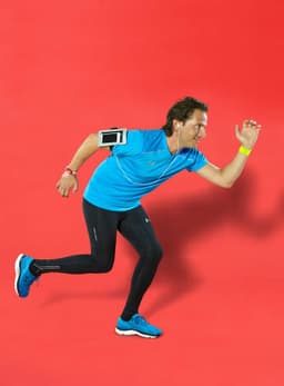 A person in a blue shirt and black leggings runs against a red background, wearing a silicone arm strap with a device.