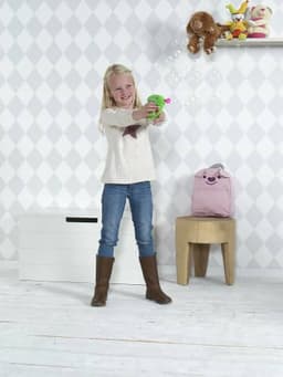 A green plastic bubble gun is being used by a child in a light-coloured room with toys and a backpack in the background.